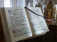 Noten der Kiedricher Chorbuben Altes Gesangsbuch der Kiedricher Chorbuben mit Noten in Hufnagelnotation und Texten in Latein in schwarz und rot gezeichnet. Im Hintergrund ist der Altar und der goldene Hochaltar zu sehen.