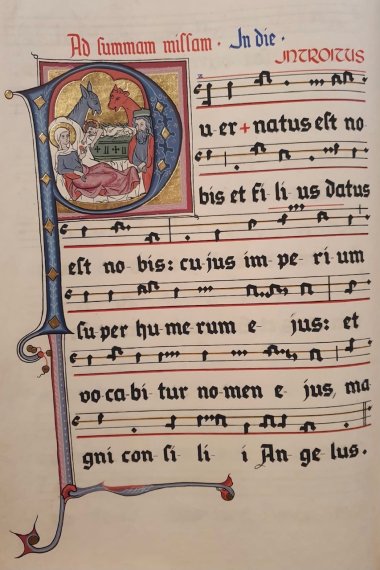 Eine Seite des alten Gesangsbuches der Kiedricher Chorbuben mit Noten in Hufnagelnotation und Texten in Latein in schwarz und rot gezeichnet. Zu Beginn ist in einem "P" in blau mit goldenen Verzierung Maria und Josef und das Jesukind in der Grippe zusammen mit dem Esel und dem Ochsen gezeichnet. 