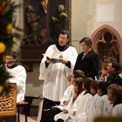 In der Kirche sitzen acht Kinder mit weißen Chorröcken, dahinter Zuschauer. Links sitzt ein Mann mit Chorrock an der Orgel mit Holzschnitzerei. Rechts davon steht Andreas Scholl im Chorrock mit einem aufgeschlagenem Buch in der Hand und singt. Er steht neben seiner Schwester Elisabeth Scholl.
