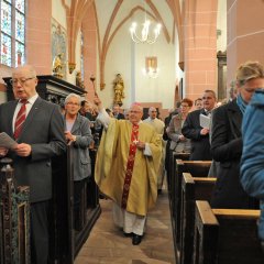 BIschof und der Pfarrer laufen durch den Seitengang der Kirche. Der Bischof weiht mit Weihrauch mit einem Stab mit einer Kugel in der Hand. In den voll besetzten Bänken stehen die Kirchenbesucher.
