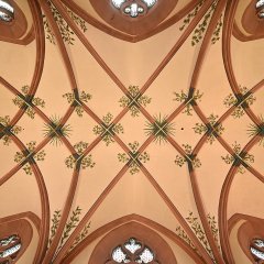 Blick an die beige Decke, an der mehrere Bögen aus Sandstein verlaufen. Zwischen den Bögen ist die Decke mit grünen Blättern, Streifen verziert. Der Kopf und die ans Kreuz genagelten Hände von Jesu sind am linken Bildrand.