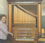 Eine ältere frau, Wilma Scholl, mit kurzen braun gelockten Haaren und einer Brille steht an einer Orgel mit Orgelpfeifen aus Holz.