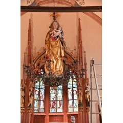 Eine Madonnafigur, die Maria hängt an der Decke der Michaelskapelle. Sie hat lange, blonde, lockige Haare und ein braunes Tuch über den Haaren und ein gold-rotes Gewand an. Sie hat das nackte Jesusbaby mit gelocktem Haar im Arm. Zu Füssen sind kleine Engelsfiguren, mit den Flügeln, Köpfen und braunem Haar. Im Hintergrund sind drei bunte Kirchenfenster und Türme aus Sandstein und vier Figuren mit langen goldenen und grünen Gewändern.