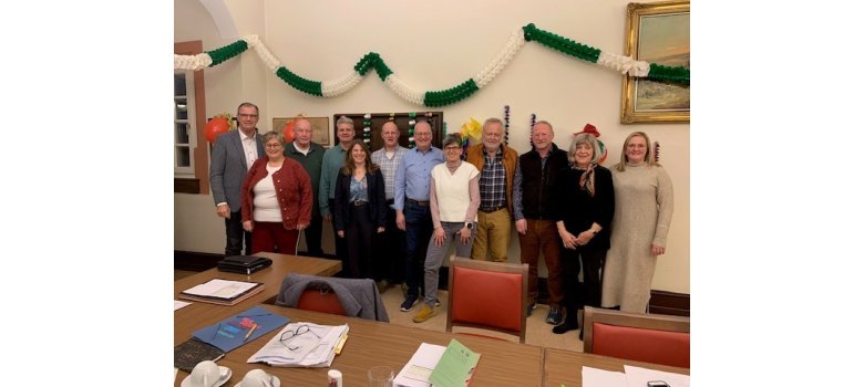 Im Sitzungssaal stehen zwölf Damen und Herren, die Mitglieder der Stuerungsgruppe nebeneinander. Im Fordergrund sind Tische und Stühle. Auf den Tischen liegen Unterlagen und Kaffee-Tassen.