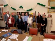 Im Sitzungssaal stehen zwölf Damen und Herren, die Mitglieder der Stuerungsgruppe nebeneinander. Im Fordergrund sind Tische und Stühle. Auf den Tischen liegen Unterlagen und Kaffee-Tassen.