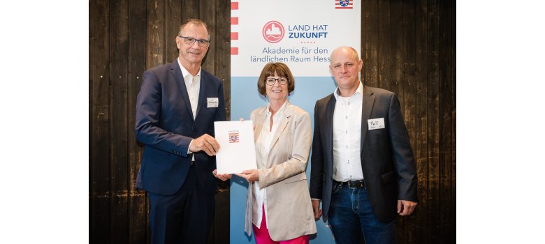 Vor einer Holzwand und einem Werbe-Banner zum Thema "Land hat Zukunft", stehen zwei Männer und eine Frau mit einer Urkundenhülle in der Hand.  