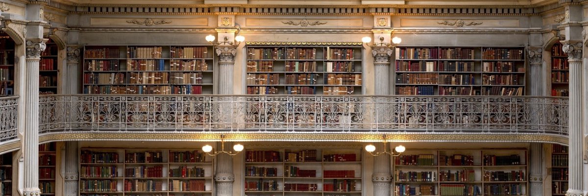 Ein Altbau mit drei Rundbögen und Gemälden, Stuck und Säulen in weiß mit Leuchten. Dazwischen sind Bücherregale und silberne Balkonbrüstungen. MAn sieht drei Ebenen.  