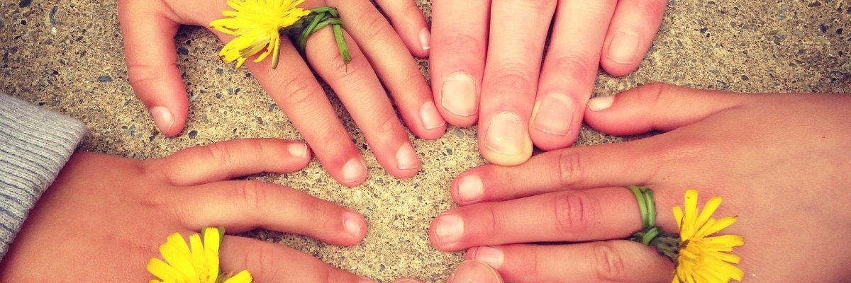 Auf einem sandfarbenem Boden sind zu einem Kreis gelegt, zwei Erwachsenen und drei Kinderhände. Jede Hand hat an einem Finger die Blüte eines Löwenzahns. Der Stengel ist um den Finger gewickelt.