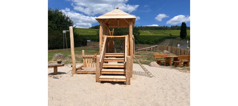 In einem großem Sandkasten steht ein Holzturm mit einer Treppe und einem Kletternetz sowie einem Zaun und einem Steuerrad. Im Hintergrund ist eine Sitzgarnitur und eine Wippe zu erkennen. Außerhalb des Zaunes sind Weinberge, oberhalb der Weinberge Wald und ein leicht bewölkter Himmel.