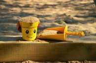 Musterbild Spielplatz Auf einem Holzbalken, vor und hinter dem sich Sand befindet, liegt eine gelbe Sandspielzeugschaufel und ein gelber Sandspielzeugeimer mit einem Gesicht und einem roten Henkel. Die Schaufel und der Eimer sind mit Sand gefüllt.
