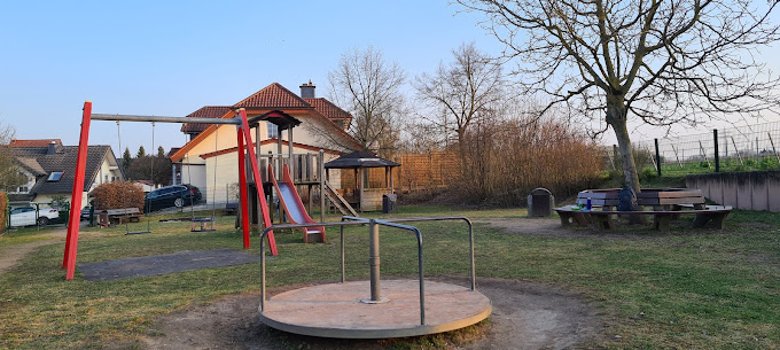 Auf einer grünen Wiese steht ein rundes Karussel, zwei Schaukeln und eine Rutsche sowie ein kleines Häuschen. Um einen Baum ist eine Sitzbank gestellt.