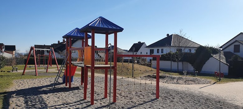 Im Hintergrund ist ein Klettergerüst und zwei Schaukeln. Davor sind zwei Türme, die mit einer Hängebrücke verbunden sind, eine Rutsche und Seile, viereckig gespannt, an denen man hochklettern kann. Drum herum ist ein Kiesbett und Wiese. 