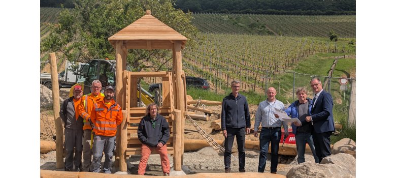 Acht Herren, darunter Bürgermeister Steinmacher, stehen vor einem Holzspielturm, mit einem Kletternetz an der Seite. Drum herum liegt weiteres Holz und Steine und Kies. Die Baustelle ist von Bauzäunen umschlossen. Im Hintergrund sind Weinberge.