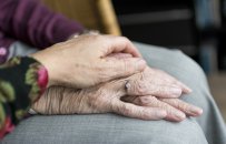 Eine Frau sitzt auf einem hellbraunen Stuhl. Sie hat eine hellgraue Hose und einen lila Pullover an. Man sieht keinen Körper und kein Gesicht.  Ihre Hände, die Falten haben, liegen aufeinander auf ihren Beinen. An dem Ringfinger steckt ein silberner Ring mit einem Stein. Eine weitere Hand, ebenfalls mit Falten, liegt augf den Händen der anderen Frau. Der Ärmel ist schwarz mit pink-roten runden Blüten, die mit runden grün-braunen Blättern umrandet sind.