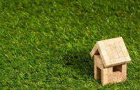 auf einer Rasenfläche steht rechts ein kleines Holzhaus, mit einer Türöffnung, einer Fenseröffnung und einem Schrägdach, welches aus Holzwürfeln errichtet wurde.