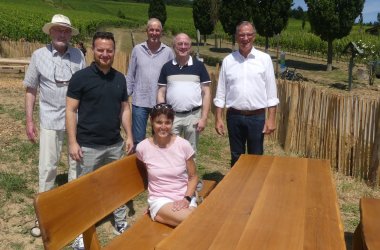 Eine Frau sitzt mit kurzer Hose und T-Shirt auf einer Bank mit Lehne an einem Holztisch. Dahinter stehen fünf Männer. Im Hintergrund ist eine Wiese und ein Holzzaun sowie Weinberge zu sehen. 
